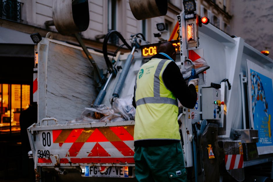 A waste collection worker wearing a high-visibility yellow vest and dark clothing operates the controls of a large, white waste disposal vehicle parked outside a building during dusk or early evening.