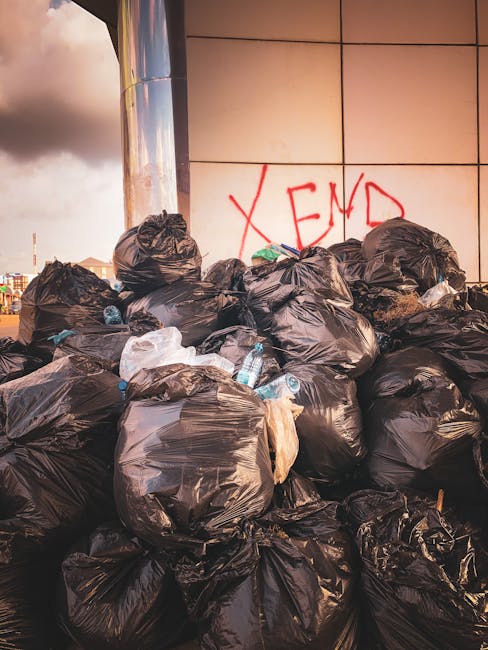 A waste collection worker wearing a high-visibility yellow vest and dark clothing operates the controls of a large, white waste disposal vehicle parked outside a building during dusk or early evening.