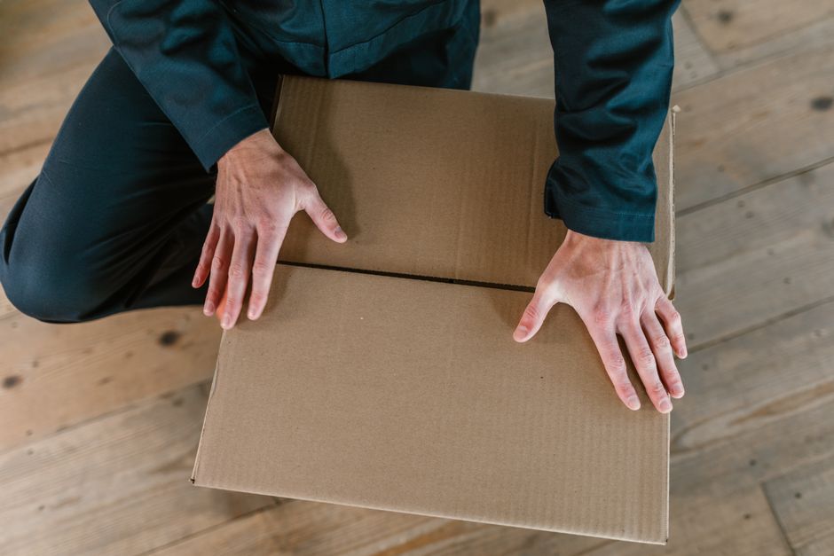 A man with curly dark hair and a beard, wearing a beige shirt, is seen lifting and carrying a medium-sized cardboard box labeled 'OFFICE' with black tape and a white label, during an office relocation