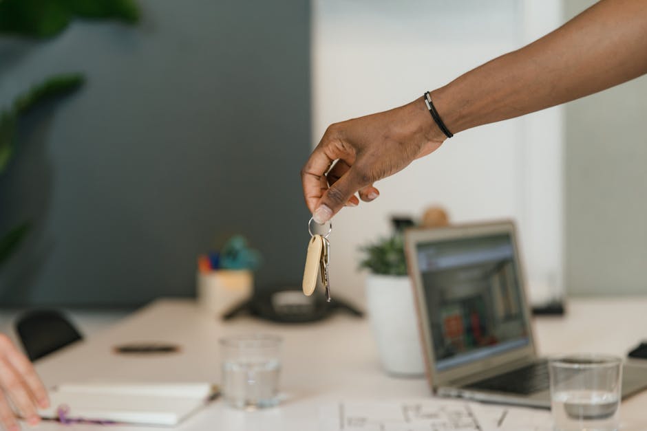 A person’s hand is extended to receive a small metal key on a keyring from another individual’s hand, which is positioned above it. The background is a plain, light grey wall, and the scene appear