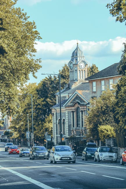 A street scene with a historic church featuring a tall clock tower and a pointed spire, set against a bright blue sky with some clouds. In the foreground, several cars are parked along the pavement, w